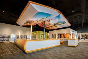 The two book-shaped shelving sections are millwork wrapped with 3M cast vinyl on the tops, sides and returns; the “pages” are 3M calendared vinyl mounted to risers and printed to match the upper canopy pages. The U-shaped units enclose a book storage area positioned beneath the lively graphics of the hapless coyote  in the wild. Photos: Moss Inc.