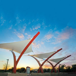 Commuters on the metro transit bus system in Mesa, Ariz., can appreciate the relief from the sun provided by the West Mesa Park and Ride. A dynamic crisscrossed row of curved red steel masts support the white fabric shade canopies over the bus passenger waiting platform. Detail: Stainless steel clevis ends connect the catenary edge cables to the painted steel supports, a welded fabric “clamping plate” in the center gathers the forces from the saddle-shape fabric panels, and a stainless toggle tensioning fitting attaches to the support. Fabric is PTFE-coated fiberglass (Sheerfill V from Saint Gobain Performance Plastics); stainless steel fittings are from Ronstan Tensile Architecture. Photo: Eide Industries Inc.
