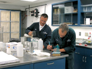 Matthew Logan (left), product application engineer at APV Engineered Coatings, closes a gallon pail with a mallet, while Gregory Marty, chemist and QC manager, tests Pencil Hardness on a sample of coated material using graphite pencils. The test allows him to evaluate the value of hardness of a particular coating formulation. Photo: APV Engineered Coatings. 
