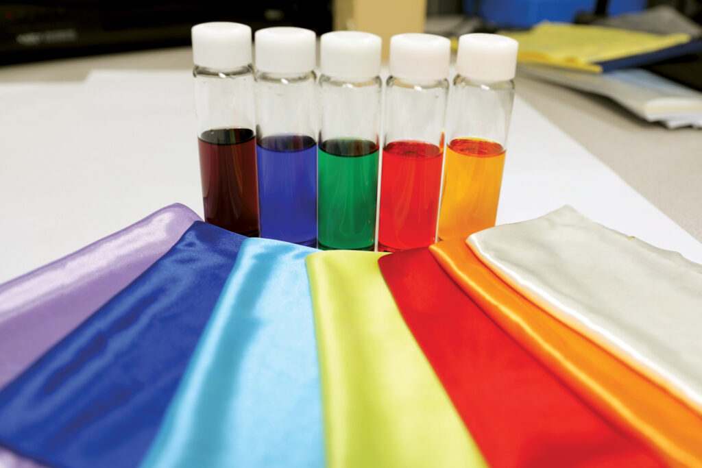 Glass vials display extracted liquid solutions containing recycled dyes derived from waste polyesters, while in front of these vials are colored polyester textiles made from recycled materials.