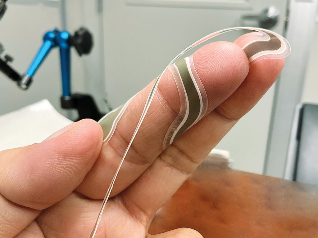 A thin, flexible heater shown wrapped around a person's finger. The heaters can be heat-pressed onto fabric to create smart textiles.