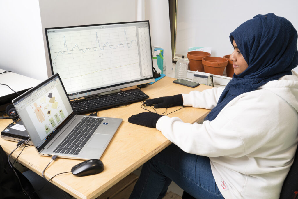 University of the West of England Bristol doctoral student Marzia Dulal tests the sensing properties of the smart gloves. She has her hands flat on the desk and is looking at a laptop that is also connected to an external monitor.