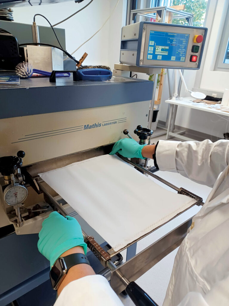 Drying/fixing a sample of polyester/lyocell fabric in the DITF laboratory tenter frame. The antimicrobial fabric is finished with AGXX. The researcher is wearing a white lab coat and medical gloves.