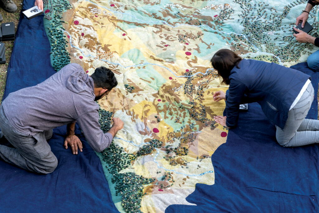 A group of artists working on a quilt. One person works on the left side of the quilt and another works on the right side.
