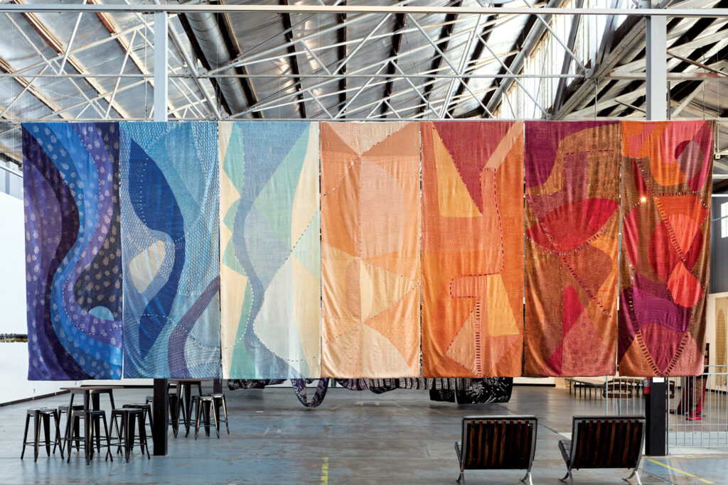 Seven large-scale, colorful textile pieces by Morii Design hanging at an event in 2024. Colors of pieces from left: dark and light blue, light blue, white and light blue, peach, orange and red, orange-red and deeper red, maroon, red and orange.