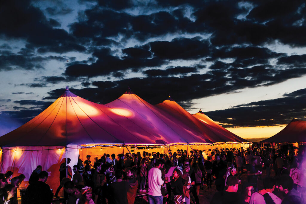 A lit up tent with purple and orange colored lights at a musical festival at sunset. Festival-goers surround the tent.