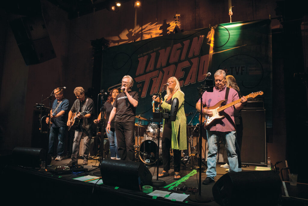 Hangin' By A Thread is a cover band made up of textile industry professionals that performs annual at Advanced Textiles Expo. Seven participants are pictured on stage here, three with guitars, the lead singer in the middle and one with a tambourine. Others are performing background vocals or are partially hidden. Five men and two women are on stage.