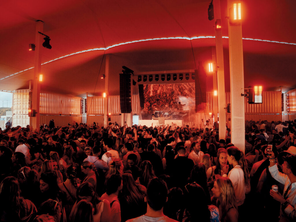 The inside of a tent at a musical festival. The lighting appears to be orange. A crowd of people watch the artist perform on stage.