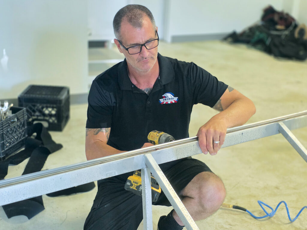 Devin Thompson installing C-rail into the top bar of an awning frame. He is kneeling on a concrete floor and using a drill.