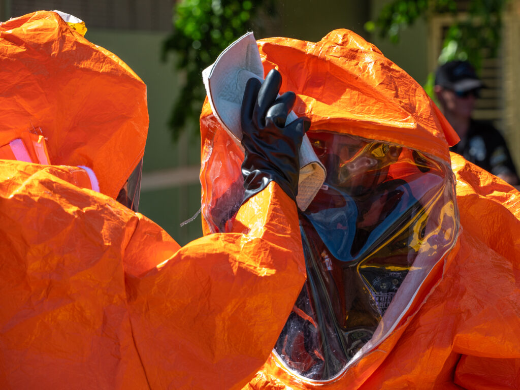 A District of Columbia 33rd Civil Support Team member simulates decontaminating another member during a field exercise. They are dressed in orang hazmat gear. Decontamination is being done with a FiberTect dry decontamination wipe.