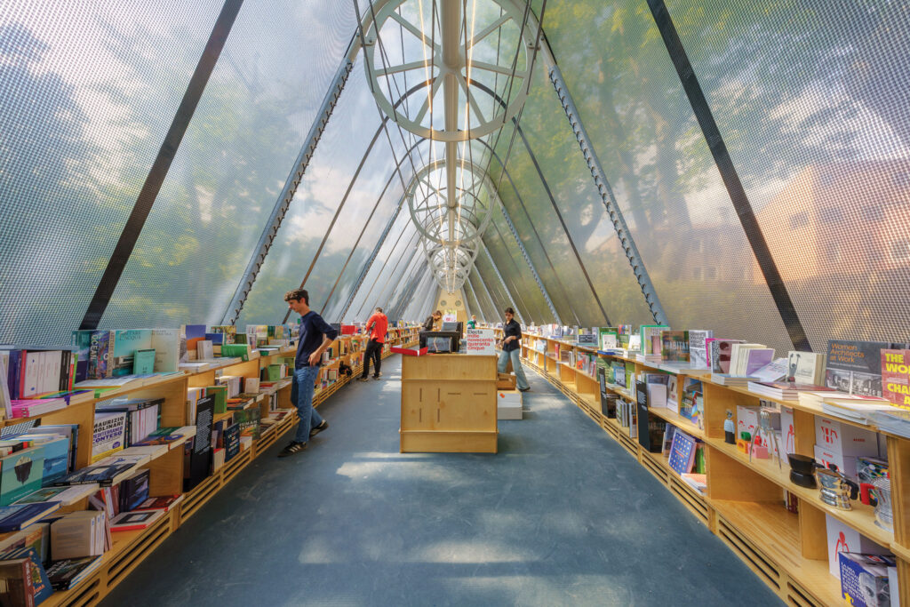 a fabric architecture bookstore with a blue floor and tan bookshelves on either side filled with multicolored books. On the left, a person stands, browsing the books.. The walls are transparent and there are trees visible.