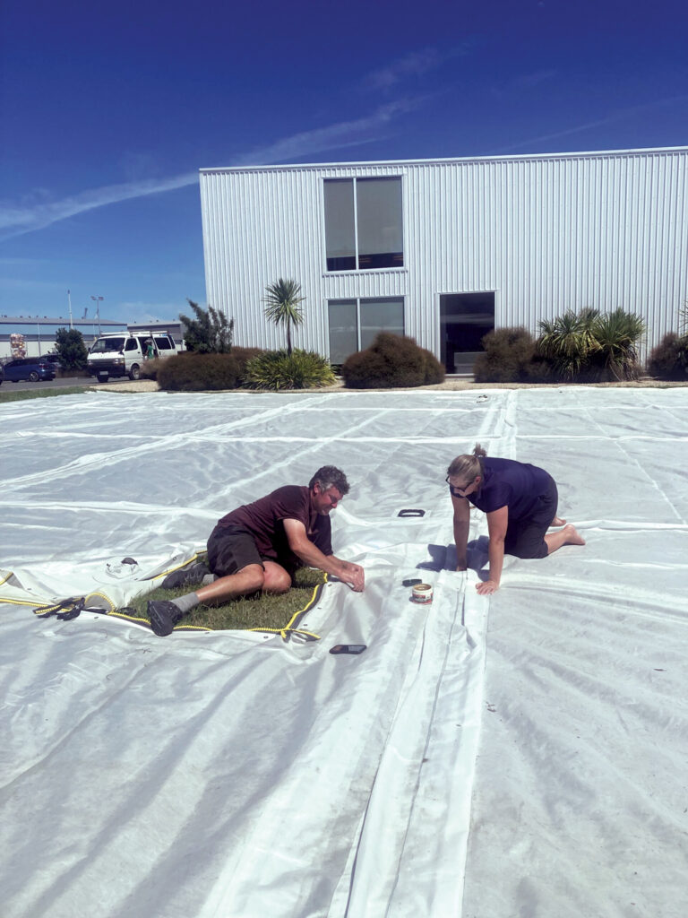 The manufacturing of the mobile fish farm. Two people are working on the giant fabric item, which is spread out end to end across the yard and extends beyond the photo