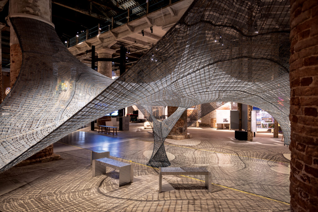 A large gray knited textile sculpture whose design and the lighting of the room result in various lines of shadows on the brown flooring. There are two silver benches underneath the sculpture.