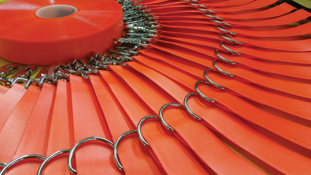 Many orange coated polyester webbing strips with D-rings and fasteners at one end arranged in a circle around a roll of orange webbing