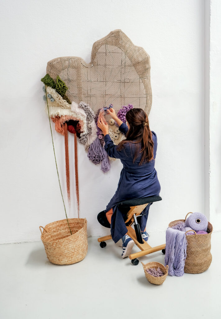 A woman works on a textile art piece installed on a white wall.