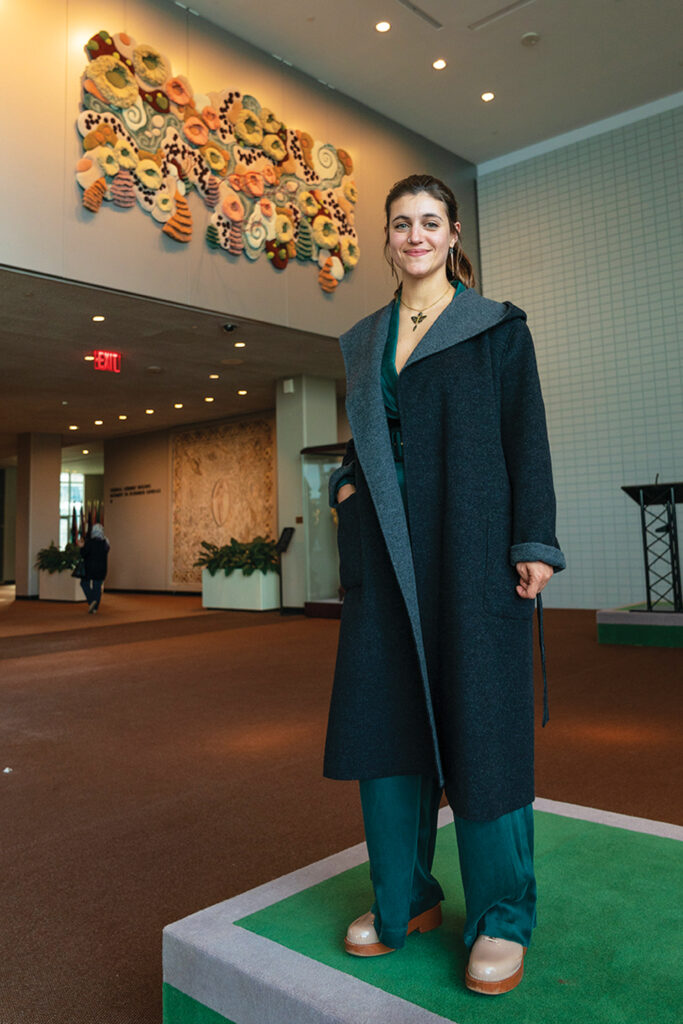 A person wearing a dark coat and teal outfit stands on a platform in a hallway with warm lighting. A colorful abstract wall art piece is displayed above.