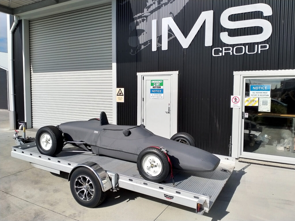 The gray custom car cover shown fully on the trailered race car in front of the IMS Group building. The travel cover fits tightly around the car and zips closed on top of the seat.