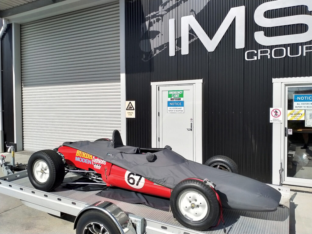 The race car on a trailer showing how the custom cover goes on the car. A portion of the lower half of the car is showing. It is red and has the number 67 on it. It is in front of a building that says IMS Group.