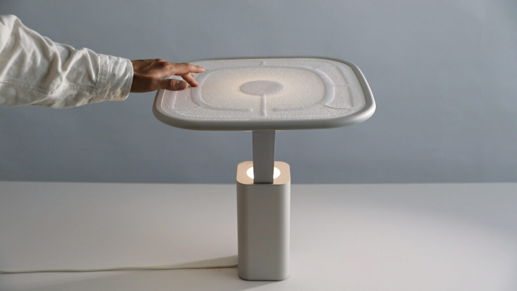 A person's finger touching the raised area of the tensioned textile on the top of the lamp. The lamp is shaped like a T, with the tensioned textile at the top horizontally. 