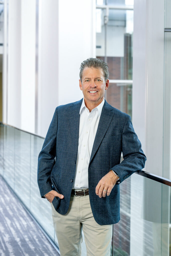 Craig Zola, new Advanced Textiles Association chairman of the board, in a dark blazer and white shirt stands confidently by a glass railing, with bright, modern architectural elements in the background.