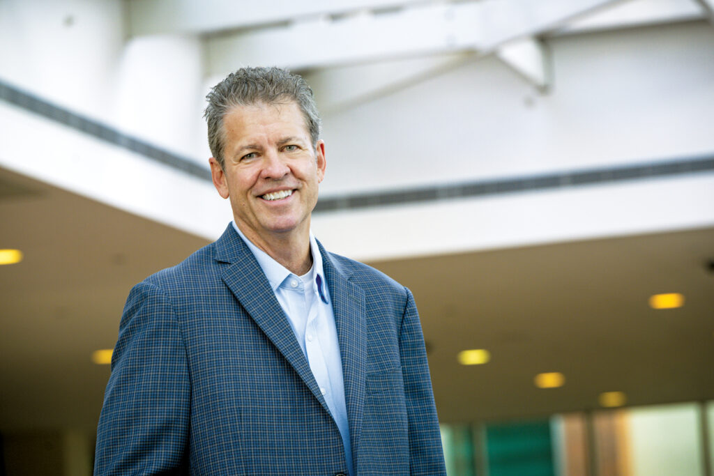 Craig Zola, new Advanced Textiles Association chairman of the board, in a blue checkered suit standing and smiling, with modern architecture and soft lighting in the background.