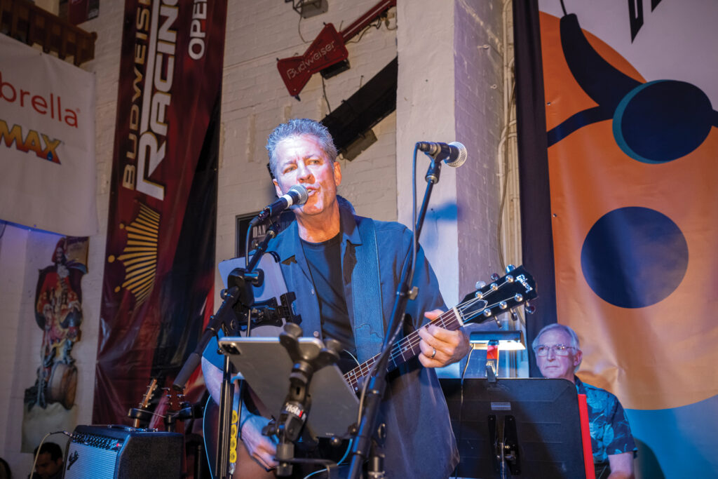 Craig Zola, new Advanced Textiles Association chairman of the board, playing guitar onstage with Hangin' by a Thread, at Advanced Textiles Expo, with a microphone and tablet stand, colorful banners and lights in the background.
