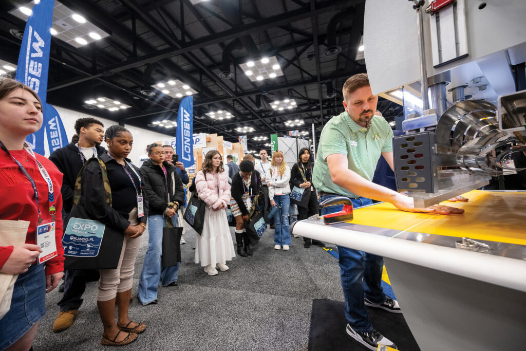 A man from Miller Weldmaster demonstrates an RFlex radio-frequency welding machine to an engaged audience of Ball State University students visiting Advanced Textiles Expo.