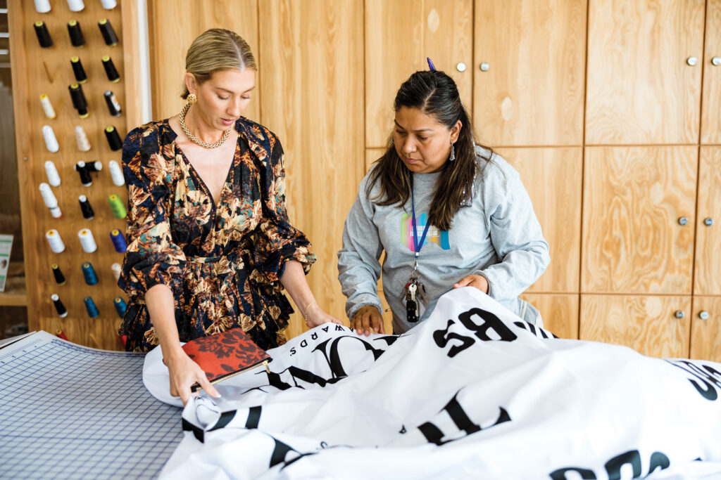 Two women collaborate on a large fabric piece, with one examining a part while the other assists, surrounded by wooden cabinetry and spools of thread.