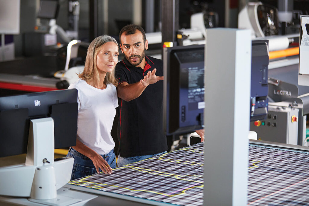 Two professionals, a man and a woman, engage in a discussion by computers in a workshop with vibrant textile patterns in the background.