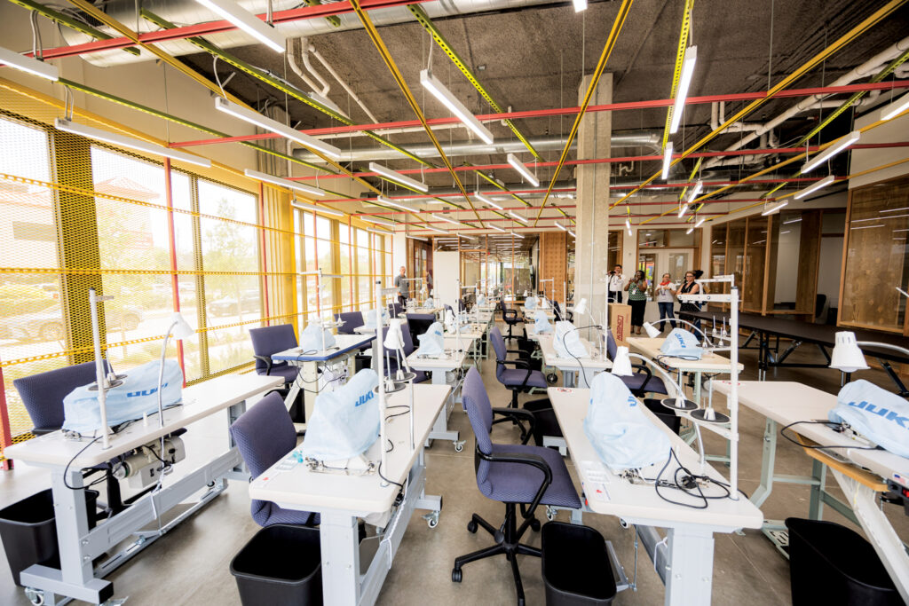 Bright, spacious workshop with sewing machines at white desks, colorful overhead cables, and large windows allowing natural light in.