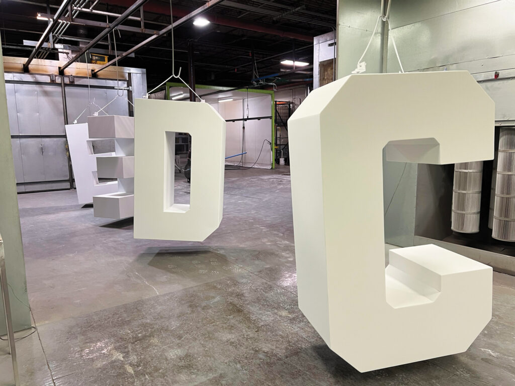 Large, white, metal letters "E," "D," and "C" are suspended in a well-lit industrial space at Reed Powder Coating to dry, with shelving and equipment in the background.