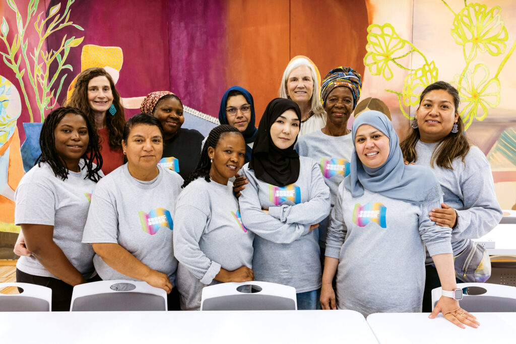 A group of women wearing gray shirts, smiling and posing together in a colorful setting with abstract wall art.