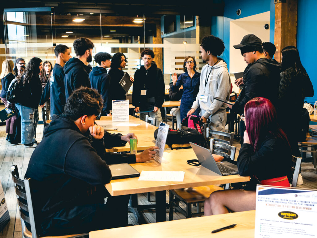 A group of young individuals network in a bright, modern space, with some seated at tables using laptops and others engaged in conversation.