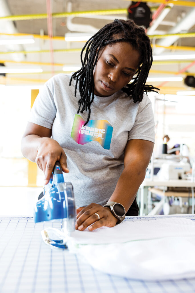 A woman with long black hair irons a piece of fabric on a blue checkered table, focused on her task in a bright workshop. She wears a gray shirt that says "Fabric Forge."