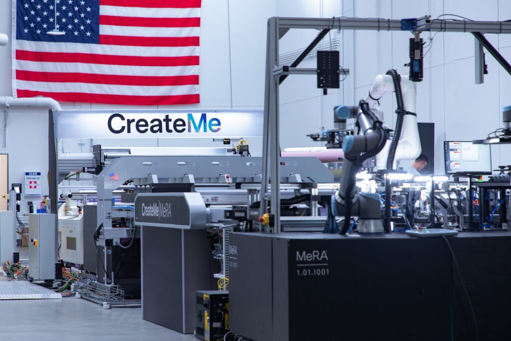 Automated textile manufacturing setup featuring advanced machinery and robotics, with an American flag in the background and "CreateMe" signage. It's no-sew automation.