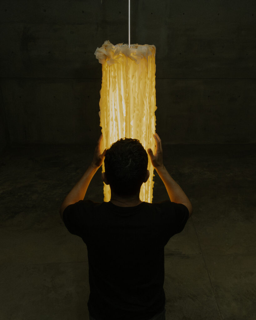 A person in a black shirt holds a glowing cylindrical lamp made from textured yellow material, set against a dark concrete background.