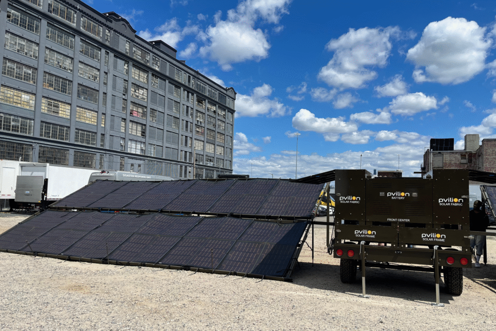 Solar panels are angled on gravel, with a dark trailer labeled "pvilion" nearby. A grey building and blue sky with clouds provide the backdrop.