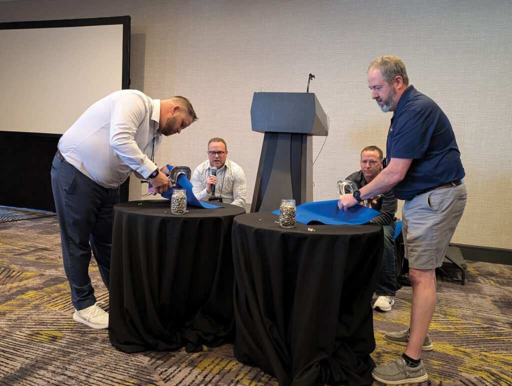 Two men interact with blue material on black tables in a conference room, while two others observe and engage from behind.