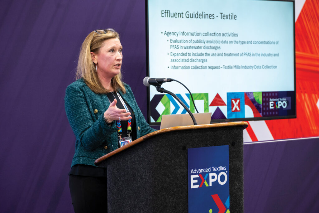 Amy Berg presents at a Advanced Textiles Expo in 2025, speaking about PFAS-related guidelines and proposals, with a colorful background and presentation screen behind her.