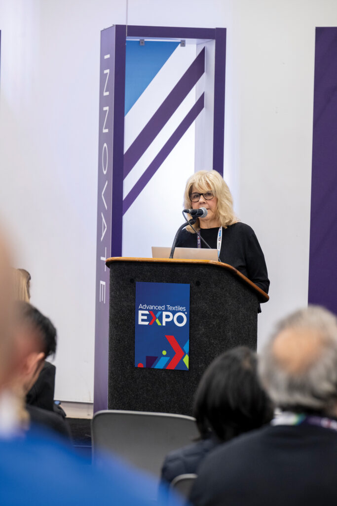 Elizabeth Denly speaks at Advanced Textiles Expo 2025 on PFAS, addressing an audience in front of a purple backdrop featuring geometric patterns.