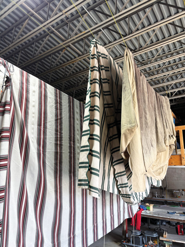 Fabric awnings in various colors, including striped and solid beige, hang from a ceiling in a workshop space with tools in the background. Al's Awning Shop provides cleaning services to its customers.