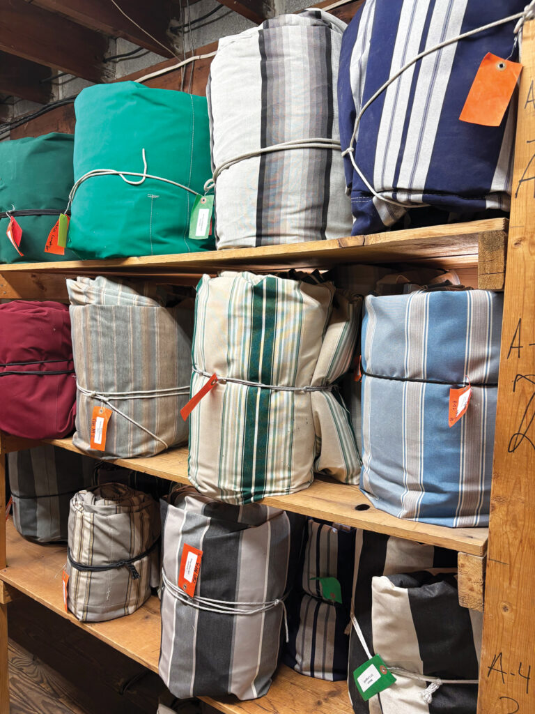 Awnings rolled and wrapped up in storage for customers of Al's Awning Shop. They are in assorted striped and solid fabrics,  tagged, and neatly stored on wooden shelves in a well-lit storage area.