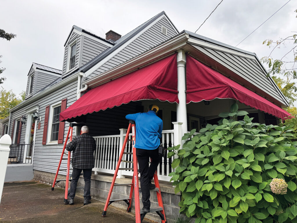 Two men on ladders from Al's Awning Shop begin taking down a red awning on a gray house with white trim and green foliage in the foreground under a cloudy sky.