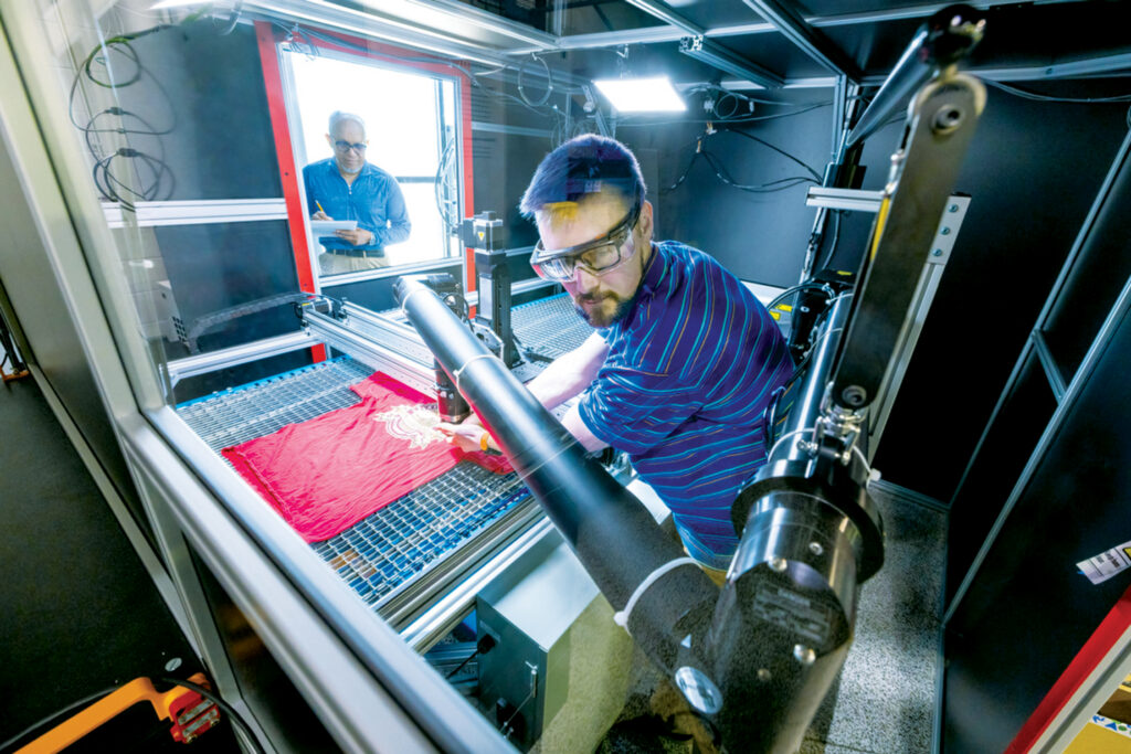 Ryan Parsons, a man in a striped blue shirt, calibrates a machine examining a red t-shirt, while Abu Islam observes from a nearby window.