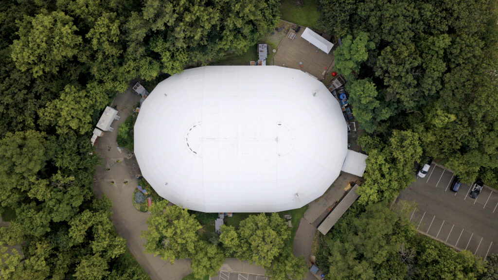 White, oval roof surrounded by dense greenery, with pathways and structures visible below, depicting a spacious outdoor area.