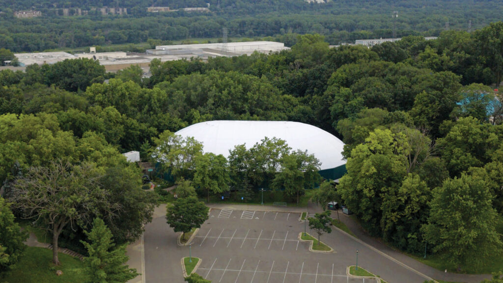 A building with a large white oval tensioned fabric roof made of Shelter-Rite material. The structure is surrounded by dense green trees, with a partially empty parking lot in the foreground.