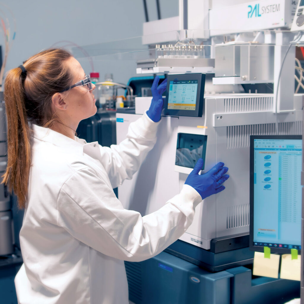 A female ClarosLabs scientist in a lab coat and gloves operates equipment, interacting with a touchscreen on a device, with a computer monitor displaying data.