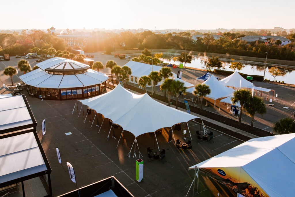 DJI_20260129204723_0776_D Spacious outdoor venue featuring white tents and a circular building, bathed in warm sunlight, near a waterway.