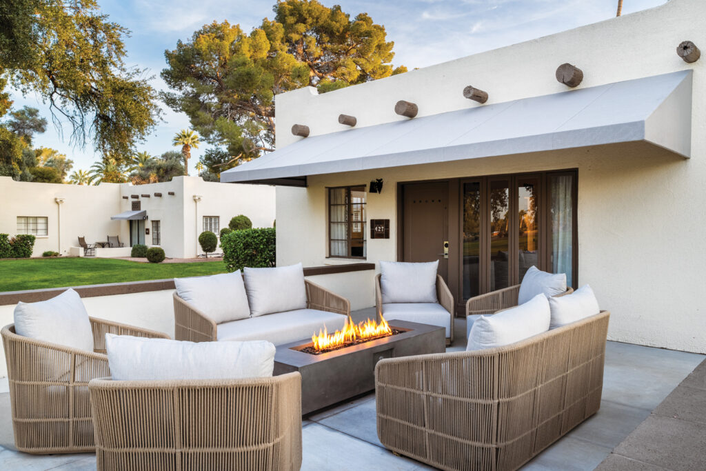 Stylish patio at resort The Wigwam, featuring a modern fire pit surrounded by beige seating, set against a backdrop of lush greenery and a white building. Th e patio is enhanced by a long light-colored stationary awning installed by Liberty Awning and Shades.