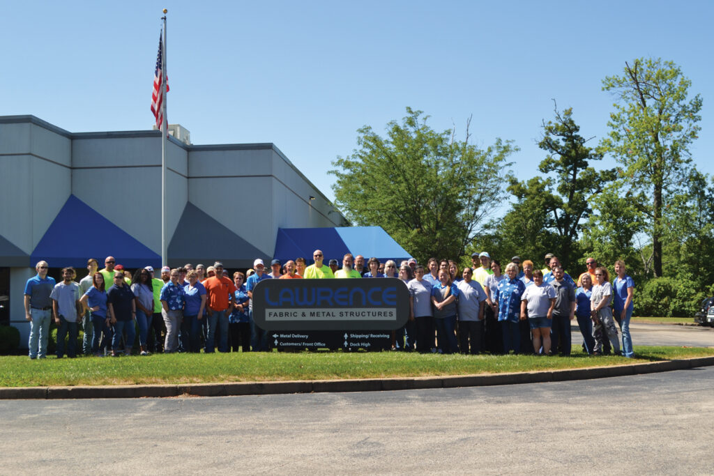 Group photo of Lawrence Fabric & Metal Structures employees outdoors, in front of a building with a blue and gray design, under a clear sky.